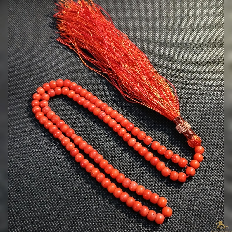 May include: A set of bright red prayer beads with a matching tassel. The beads are round and strung together, and the tassel is made of fine, red threads. The beads and tassel are displayed on a dark, textured surface.