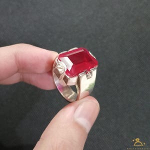 Op de afbeelding: Zilveren ring met een grote, rechthoekige, gefacetteerde rode edelsteen. De ring heeft een brede band en decoratieve elementen op de schouders, wat een gedurfd ontwerp creëert. De rode edelsteen is het middelpunt.