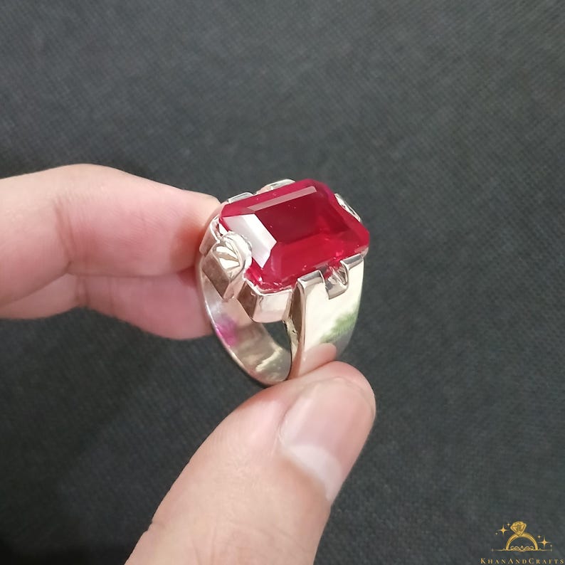 Puede incluir: Anillo de plata con una gran gema roja rectangular facetada. El anillo tiene una banda ancha y elementos decorativos en los hombros, mostrando un dise&ntilde;o audaz. La gema roja es el punto focal.