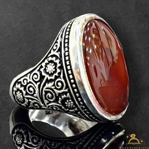 Puede incluir: Un anillo de plata con una piedra preciosa ovalada de color marrón rojizo. El anillo presenta intrincados diseños florales y de volutas en la banda. La piedra preciosa está engastada en un bisel de plata. El anillo se muestra sobre una superficie reflectante.