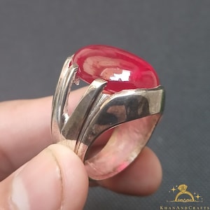May include: A silver ring featuring a large, oval-shaped, red gemstone set in the centre. The ring has a distinctive, intricate design with a silver band that wraps around the gemstone.