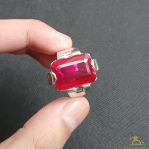 Puede incluir: Un anillo de plata con una gran gema roja rectangular y vibrante. El anillo tiene un dise&ntilde;o moderno con una banda ancha y detalles decorativos en los lados de la gema. La gema es el punto focal, captando la luz y mostrando su color intenso.
