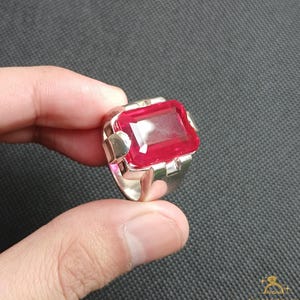 Puede incluir: Un anillo de plata con una gran gema roja brillante de forma rectangular. La gema es el punto focal, engastada en una banda de plata con detalles geom&eacute;tricos en los laterales. El anillo se muestra sobre un fondo oscuro.