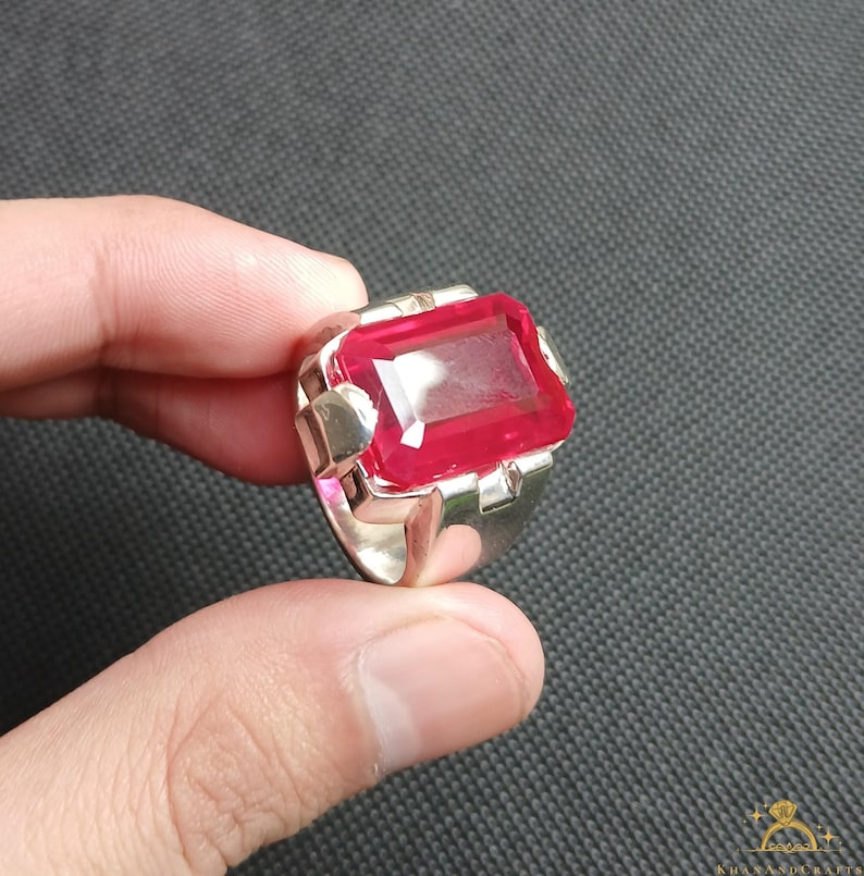 Puede incluir: Un anillo de plata con una gran gema roja brillante de forma rectangular. La gema es el punto focal, engastada en una banda de plata con detalles geom&eacute;tricos en los laterales. El anillo se muestra sobre un fondo oscuro.