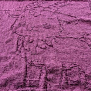 May include: A knitted blanket in a deep magenta color. The blanket features a textured design of a cartoon animal, possibly a sheep or lamb. The edges have a ribbed border.