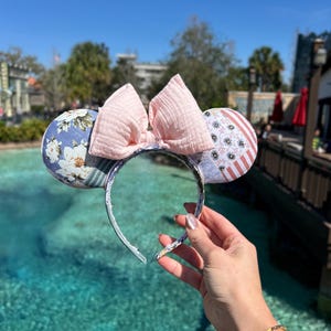 May include: A pair of pink and white Minnie Mouse ears with a floral print on one ear and a red, white, and blue print on the other ear. The ears are held in front of a blue body of water.