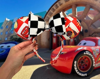 May include: Headband featuring two round ears with a red and white checkered pattern and a black and white checkered bow. The ears are decorated with images of a red race car. The headband is red and white.