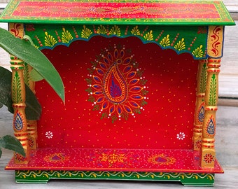Puede incluir: Un pequeño estante de madera rojo con un diseño pintado decorativo. El estante tiene un borde superior e inferior verde y detalles dorados. El estante está decorado con un patrón de paisley y puntos blancos.
