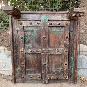 May include: A distressed wooden cabinet with two doors, metal studs, and a latch. The wood displays brown and green hues. Decorative elements are present at the top. The cabinet has a rustic, aged appearance.