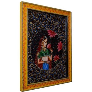 May include: A framed painting depicting a woman in a colourful sari holding a pink flower. The background is a dark blue with intricate floral patterns.