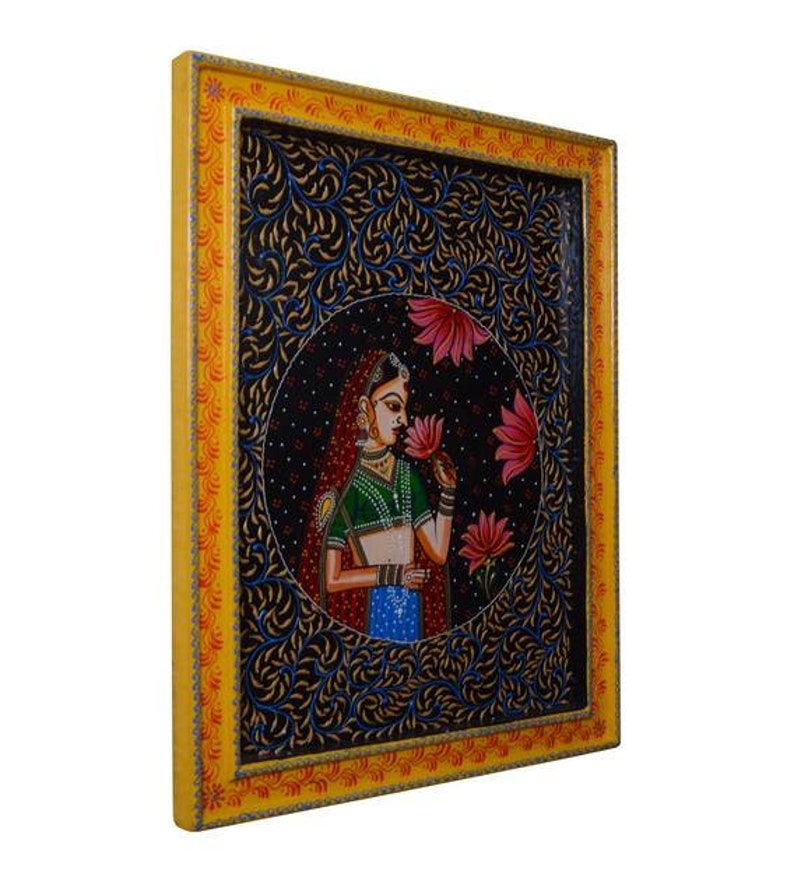 May include: A framed painting depicting a woman in a colourful sari holding a pink flower. The background is a dark blue with intricate floral patterns.