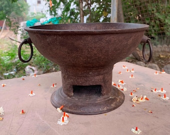 May include: A weathered, brown metal bowl-shaped object with two ring handles. The bowl sits atop a pedestal base with a rectangular opening. Small white and orange flowers are scattered around the base, suggesting an outdoor setting.