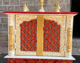 May include: A white wooden temple with red and gold accents. The temple has two doors with intricate latticework and two drawers at the bottom. The temple is decorated with floral designs.
