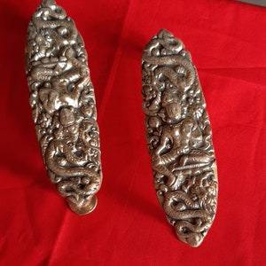 Op de afbeelding: Twee zilverkleurige, langwerpige decoratieve stukken met ingewikkelde gravures van figuren en draken. De stukken staan tegen een rode achtergrond, wat het gedetailleerde vakmanschap en ontwerp laat zien.