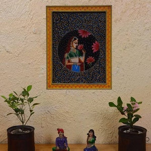 May include: A framed painting of a woman in traditional Indian clothing, holding a flower. The painting is set against a dark blue background with intricate floral patterns. The frame is gold with a yellow border.