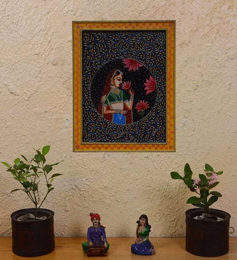 May include: A framed painting of a woman in traditional Indian clothing, holding a flower. The painting is set against a dark blue background with intricate floral patterns. The frame is gold with a yellow border.