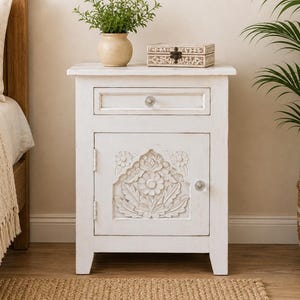 May include: A white bedside table featuring a single drawer with a clear knob and a cabinet door below. The door displays a carved floral pattern and a matching clear knob. The table has a distressed finish and stands on four straight legs.