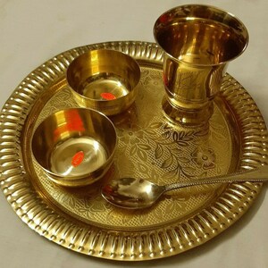 May include: A golden brass puja thali set, featuring a large round tray with a fluted edge, two small bowls, a tall glass, and a spoon. The tray and bowls have floral patterns. The set is designed for religious ceremonies.