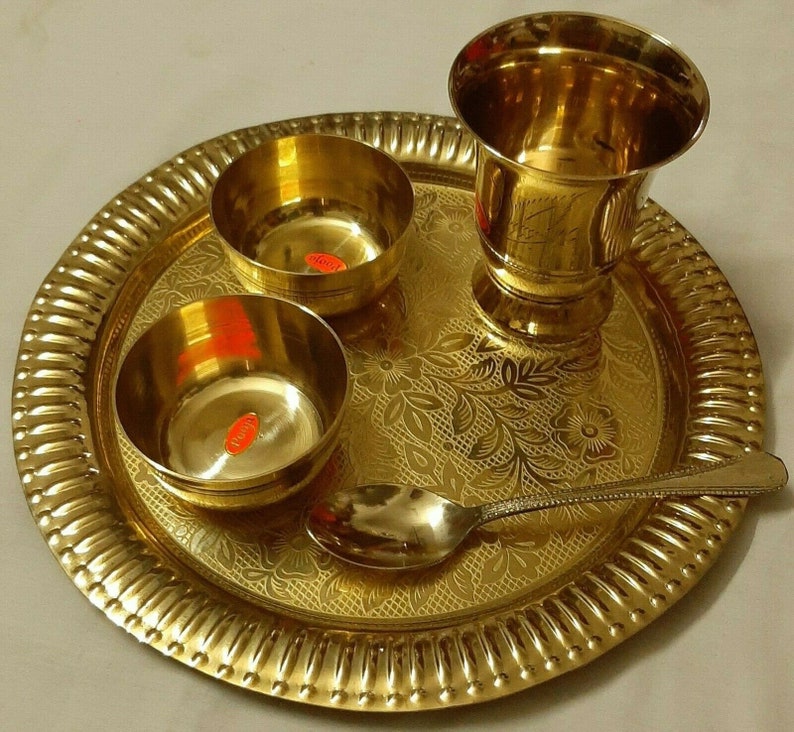 May include: A golden brass puja thali set, featuring a large round tray with a fluted edge, two small bowls, a tall glass, and a spoon. The tray and bowls have floral patterns. The set is designed for religious ceremonies.
