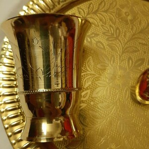 May include: A gold-colored metal cup with etched designs, sitting on a gold-colored tray with a floral pattern. The cup has a flared top and a decorative band near the middle. The tray and cup reflect light, creating a warm, inviting aesthetic.