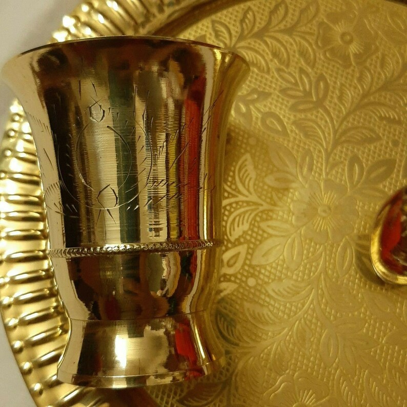 May include: A gold-colored metal cup with etched designs, sitting on a gold-colored tray with a floral pattern. The cup has a flared top and a decorative band near the middle. The tray and cup reflect light, creating a warm, inviting aesthetic.