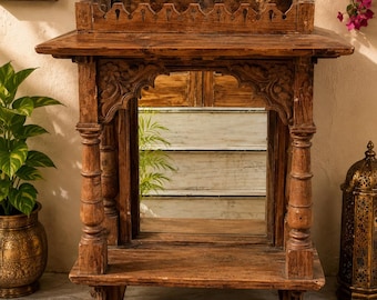 Puede incluir: Un santuario de pared de madera marrón con un espejo rectangular central, detalles tallados en arco, dos pilares torneados y un borde superior festoneado. La estructura incluye un estante plano en la base y una repisa superior decorativa.