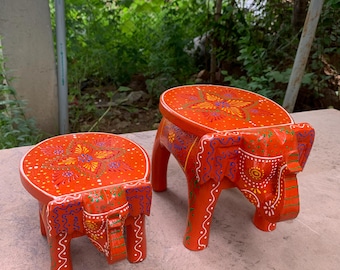 May include: Two hand-painted wooden elephant stools in orange with colorful floral designs. The larger stool is taller than the smaller stool.