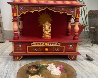 Puede incluir: Un altar de madera rojo con intrincados tallados y detalles pintados. El altar presenta una pequeña estatua de Ganesha, una deidad hindú, y un cajón con un diseño floral. El altar está decorado con flores y arroz.
