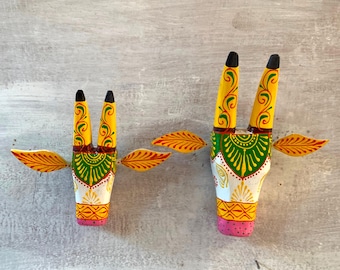 Pode incluir: Duas esculturas de cabeça de vaca de madeira pintadas de branco, amarelo, verde e rosa. A escultura maior tem um desenho floral verde e amarelo nos chifres. A escultura menor tem um desenho semelhante, mas é menor.