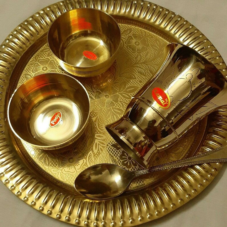May include: A golden brass serving set featuring a round tray with a floral pattern, two small bowls, a tall cup, and a spoon. The set includes labels with text. The items are arranged on a white surface.