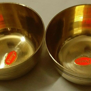 May include: Two small, round, brass bowls with a polished finish. Each bowl has a red and orange sticker with the word "Pooja" on it. The bowls are likely used for religious or decorative purposes.