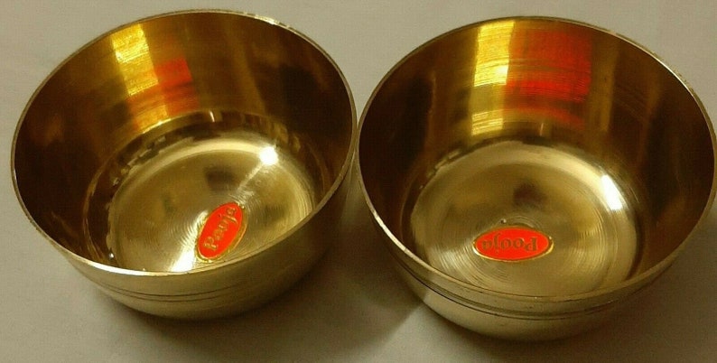 May include: Two small, round, brass bowls with a polished finish. Each bowl has a red and orange sticker with the word "Pooja" on it. The bowls are likely used for religious or decorative purposes.