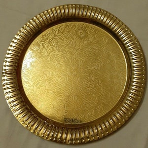May include: A round, gold-colored tray with a floral pattern on the surface. The tray has a decorative, ridged edge. The floral design is embossed on the tray's surface, creating a textured appearance.
