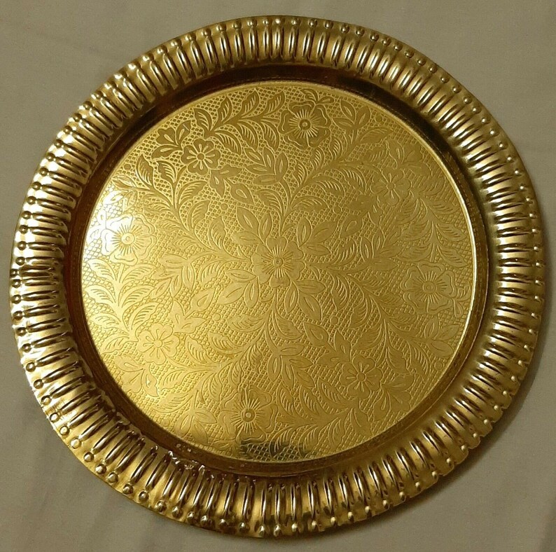 May include: A round, gold-colored tray with a floral pattern on the surface. The tray has a decorative, ridged edge. The floral design is embossed on the tray's surface, creating a textured appearance.