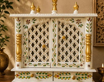 May include: A white wooden shrine featuring two doors with intricate lattice cutouts, gold-painted pillars, and decorative floral motifs in green and yellow. The structure includes two small drawers with gold knobs and four gold finials on top.
