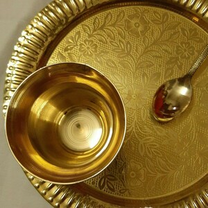May include: A gold-colored metal serving set, including a small bowl, a spoon, and a round tray. The tray has a floral pattern and a ridged edge. The bowl and spoon are smooth and reflective, creating a warm, inviting aesthetic.