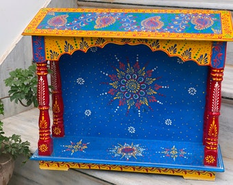 Puede incluir: Un templo de madera pintado a mano con fondo azul, ribetes amarillos y pilares rojos. El templo presenta intrincados diseños florales y un patrón de estrella.