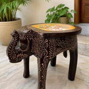 May include: A dark brown, hand-carved wooden elephant stool with a round top. The top is painted yellow with floral designs. The elephant's body and legs are intricately carved with gold accents. The stool is approximately 12 inches tall.