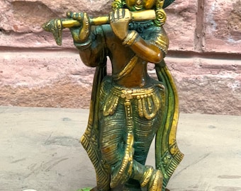 Puede incluir: Una estatua de metal dorado y verde de Krishna tocando la flauta. La estatua está de pie sobre una base redonda con intrincados detalles.