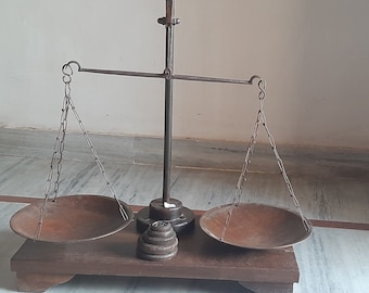 May include: An antique balance scale with two rusty metal bowls hanging from chains. The scale is made of metal and has a wooden base. The scale is used for weighing items.