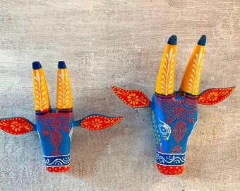 Pode incluir: Duas peças de decoração de parede de cabeça de touro de madeira pintadas. A peça maior é azul com chifres amarelos e brancos e detalhes vermelhos e laranja. A peça menor é azul com chifres amarelos e brancos e detalhes vermelhos e laranja.