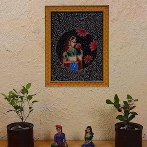 May include: A framed painting depicting a woman in traditional Indian attire, holding a flower. The painting is set against a dark blue background with intricate floral patterns. The frame is yellow and gold.