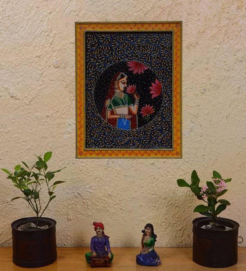 May include: A framed painting depicting a woman in traditional Indian attire, holding a flower. The painting is set against a dark blue background with intricate floral patterns. The frame is yellow and gold.