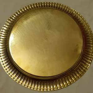 May include: A round, gold-colored metal tray. The tray has a smooth, flat center and a decorative, ridged border with small, evenly spaced indentations. The overall design is simple and elegant.