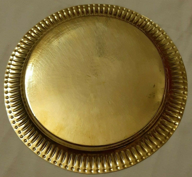 May include: A round, gold-colored metal tray. The tray has a smooth, flat center and a decorative, ridged border with small, evenly spaced indentations. The overall design is simple and elegant.