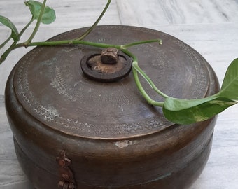 May include: A round, antique, brass box with an intricate design and a wooden knob on the lid. The box is closed and has a metal latch. A green plant is draped over the box.
