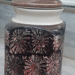 May include: A ceramic jar with a lid, featuring a black and red abstract pattern. The jar has a white rim and a brown lid.
