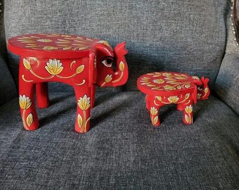 May include: Two red wooden elephant stools with gold painted floral designs. The larger stool has a round top and the smaller stool has a slightly oval top.