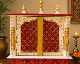 Może przedstawiać: Drewniana świątynia domowa w kolorystyce białej i czerwonej ze złotymi akcentami. Konstrukcja zawiera dwoje drzwi z czerwonym wzorem kratki, dwie szuflady z kwiatowymi detalami oraz trzy złote zwieńczenia na górnej krawędzi dachu.