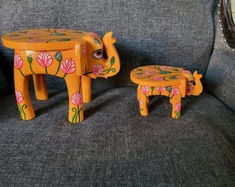 May include: Two hand-painted wooden elephant stools with floral designs. The larger stool is orange with pink flowers and green leaves. The smaller stool is orange with pink flowers and green leaves. Both stools have a round top.